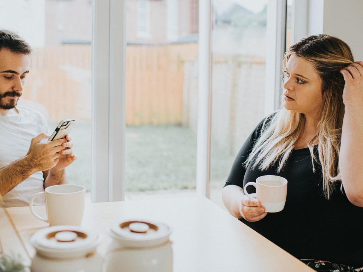 Ein Mann sitzt am Handy und seine Frau sitzt mit einer Tasse gegenüber und guckt ihn an.