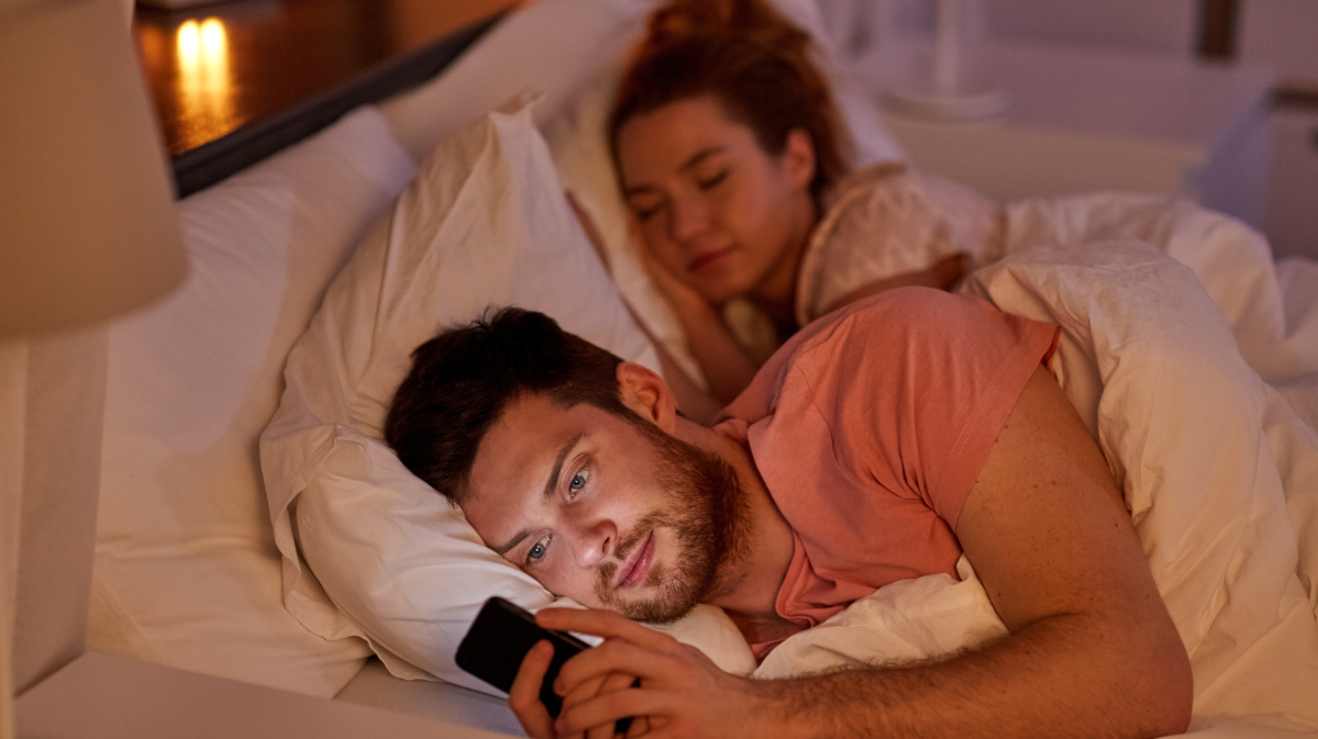 Mann, der im Bett an seinem Handy spielt, während im Hintergrund eine Frau im Bett schläft