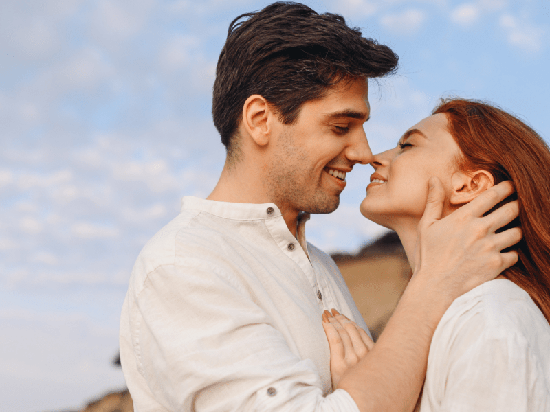 Ein Mann mit dunklem Haar und eine Frau mit roten langen Haaren stehen lächelnd nah beieinander unter klarem Himmel.
