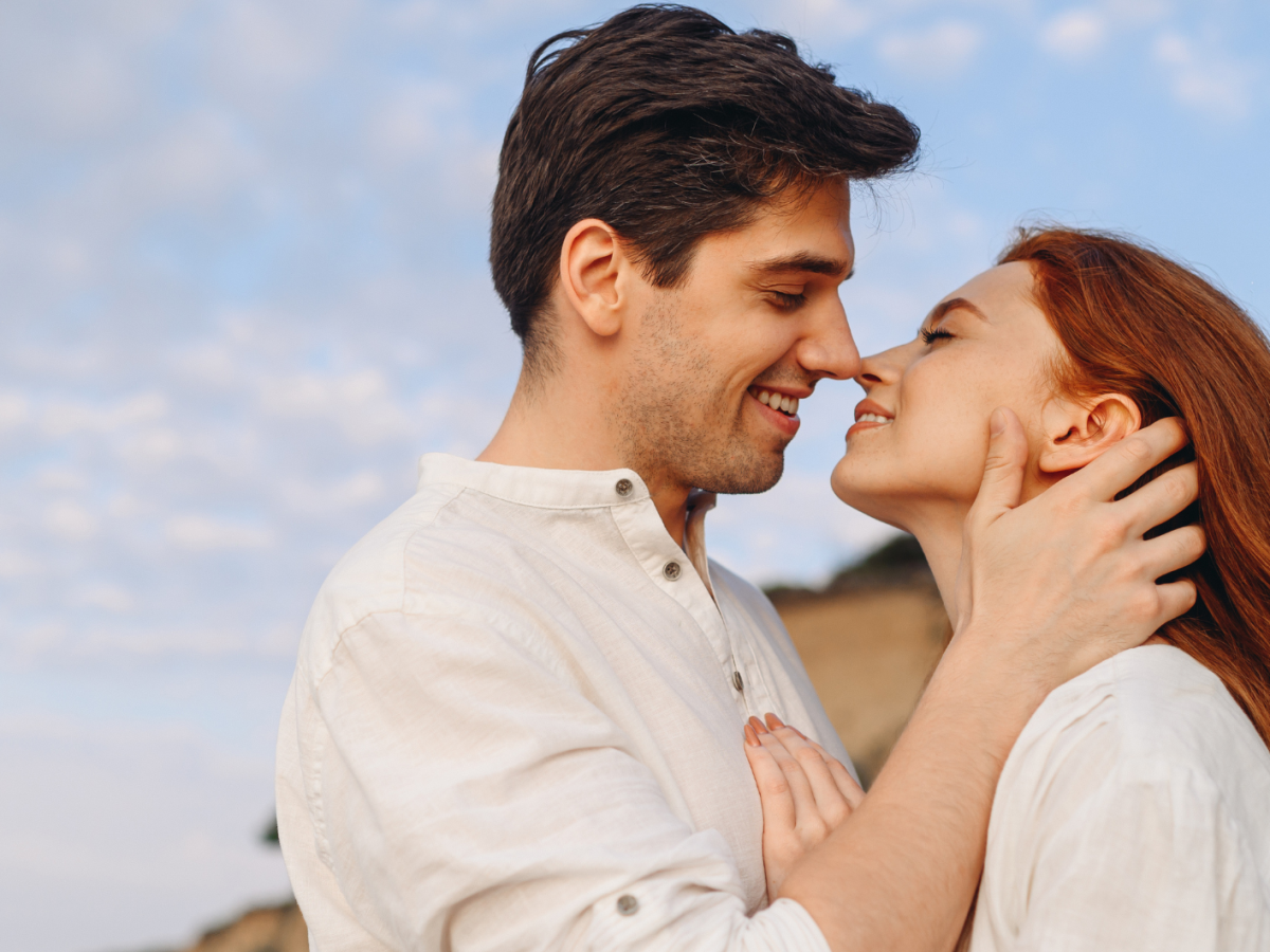 Ein Mann mit dunklem Haar und eine Frau mit roten langen Haaren stehen l&auml;chelnd nah beieinander unter klarem Himmel.