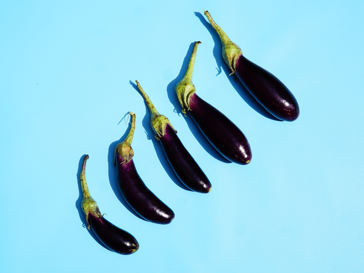 Fünf längliche lilafarbene Auberginen mit grünen Stielen liegen in einer diagonalen Linie auf hellblauem Hintergrund.