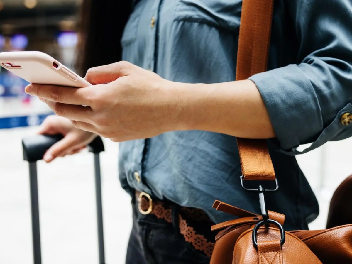 Frau am Flughafen mit einem Koffer und einem Handy und einer Reisetasche.