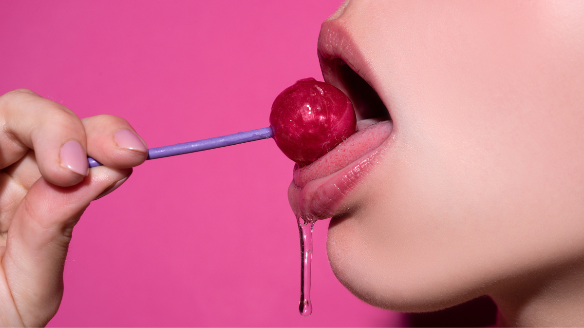 Nahaufnahme eines geöffneten Mundes mit glänzenden Lippen, der einen roten Lolli am lilafarbenen Stiel leckt, vor pinkem Hintergrund.