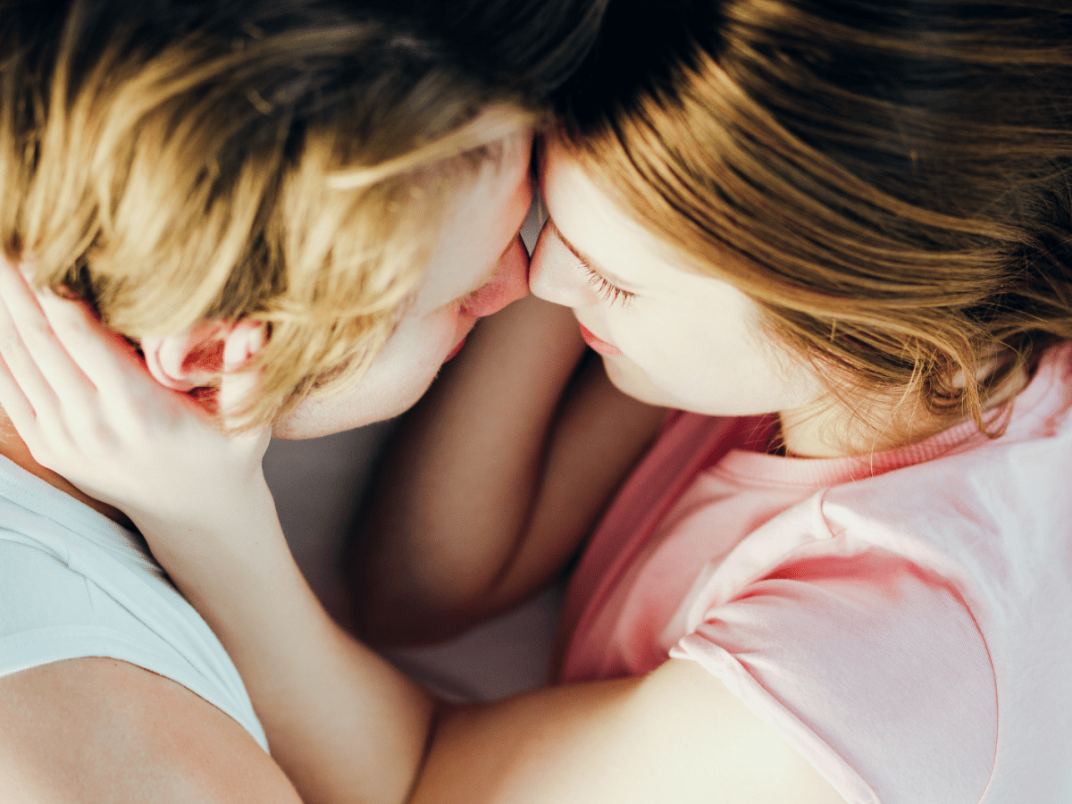 Junges Paar mit blonden Haaren liegt eng umschlungen, ihre Stirnen aneinander, sie l&auml;cheln z&auml;rtlich auf wei&szlig;em Bett.