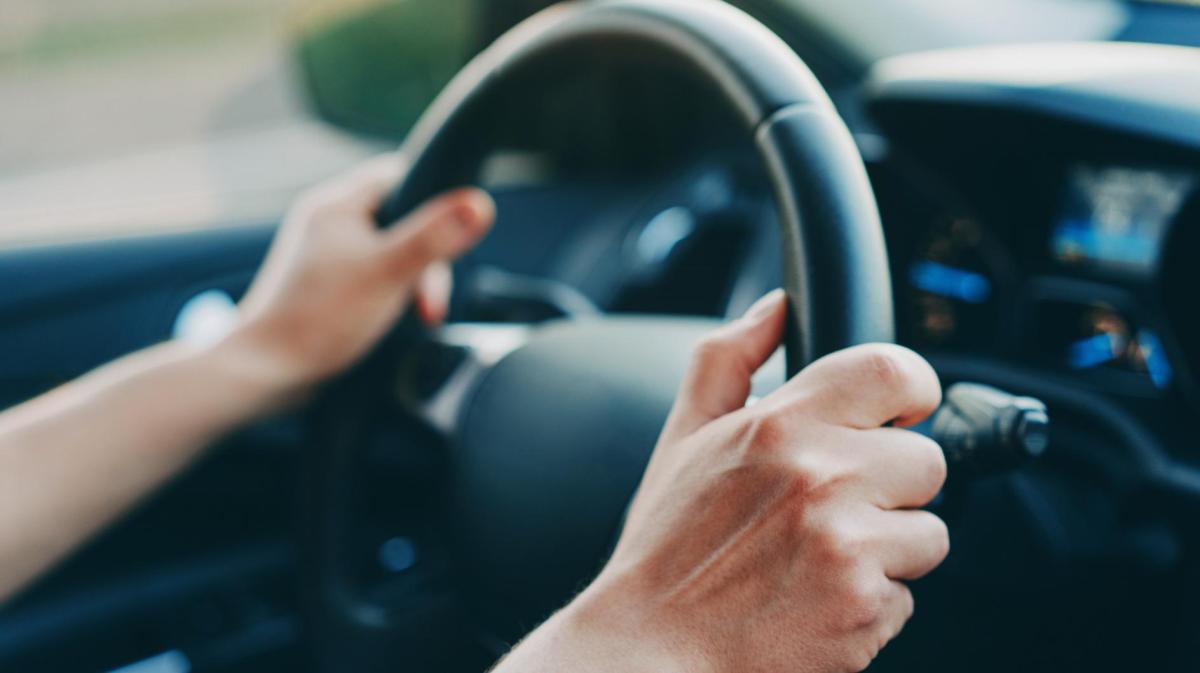 Nahaufnahme zweier Hände am Lenkrad eines Autos, mit unscharfem Armaturenbrett und Straßenhintergrund im Abendlicht.