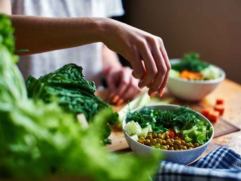 Weibliche Hand gibt gr&uuml;ne Zwiebeln in eine Sch&uuml;ssel mit gr&uuml;nen Erbsen, Gurken, Karotten, Salat und Dill, die auf einem Tisch stehen