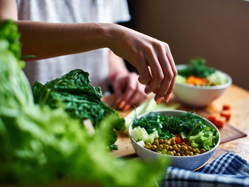 Weibliche Hand gibt grüne Zwiebeln in eine Schüssel mit grünen Erbsen, Gurken, Karotten, Salat und Dill, die auf einem Tisch stehen