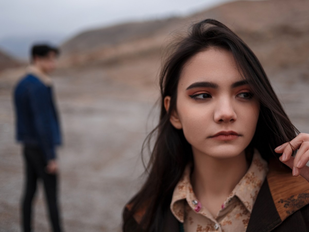 Frau in der Natur, die nach vorne schaut, während im Hintergrund ein Mann zu sehen ist, der verschwommen zu ihr rüberschaut