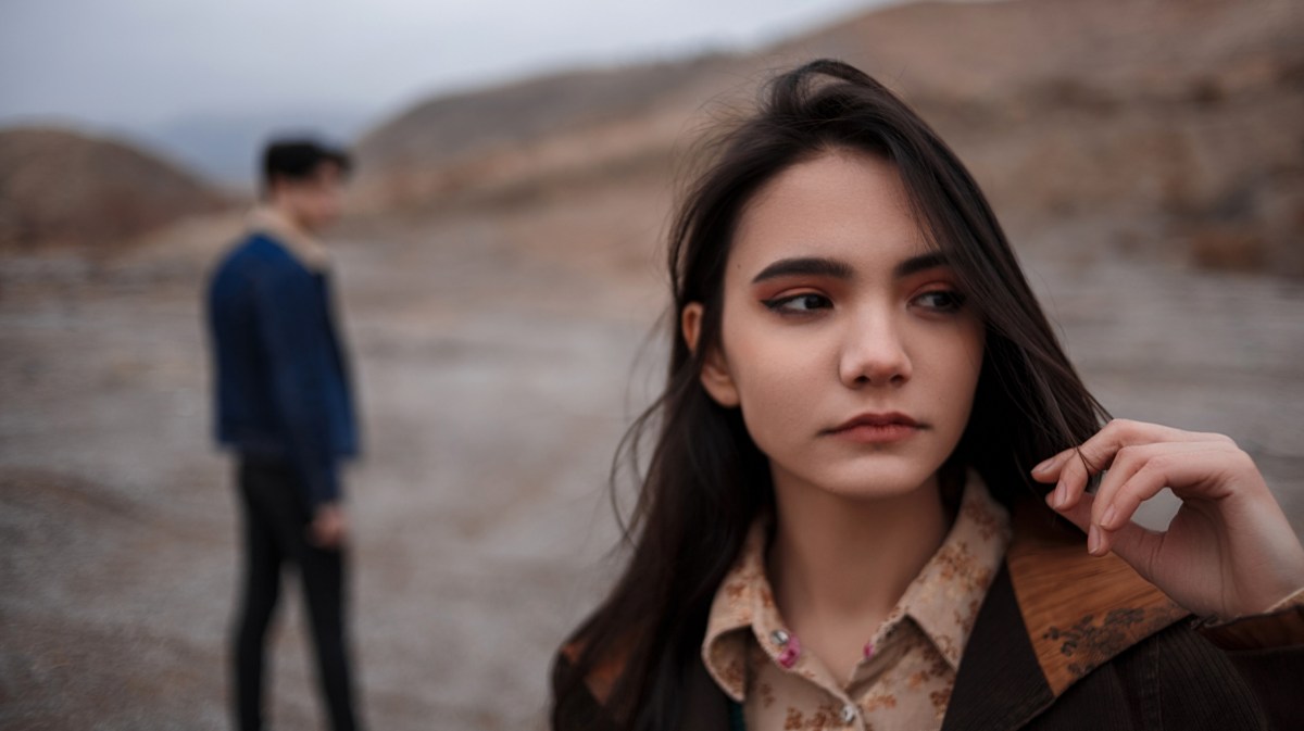 Frau in der Natur, die nach vorne schaut, während im Hintergrund ein Mann zu sehen ist, der verschwommen zu ihr rüberschaut