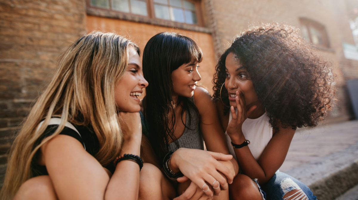 Drei Freundinnen im Sommer, die auf dem Bordstein sitzen und sich unterhalten und lachen.