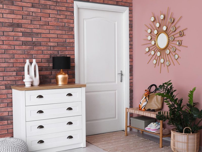 Heller Flur mit weißer Kommode, goldener Lampe und Deko-Vasen vor roter Backsteinwand neben rosa Wand mit Sonnenspiegel.