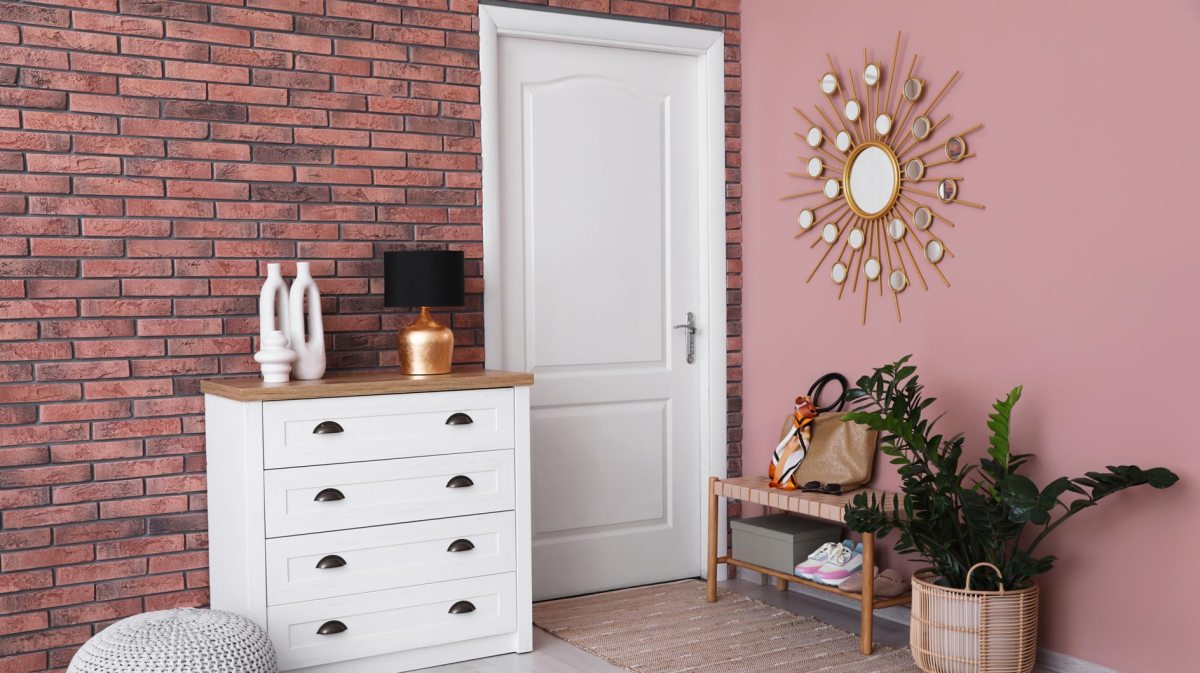 Heller Flur mit weißer Kommode, goldener Lampe und Deko-Vasen vor roter Backsteinwand neben rosa Wand mit Sonnenspiegel.