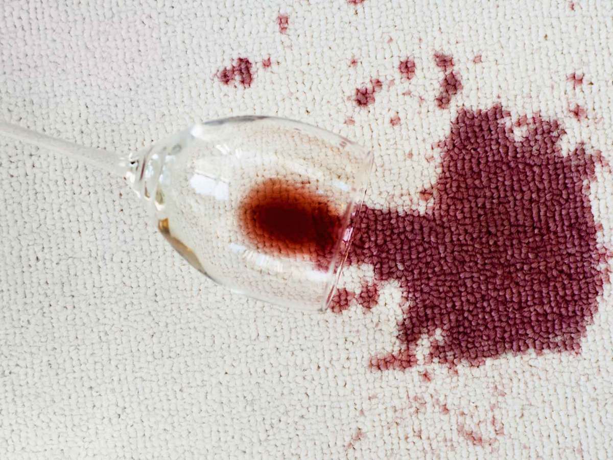 Umgekipptes Rotweinglas liegt auf einem hellen Teppich mit einem gro&szlig;en Rotweinfleck.