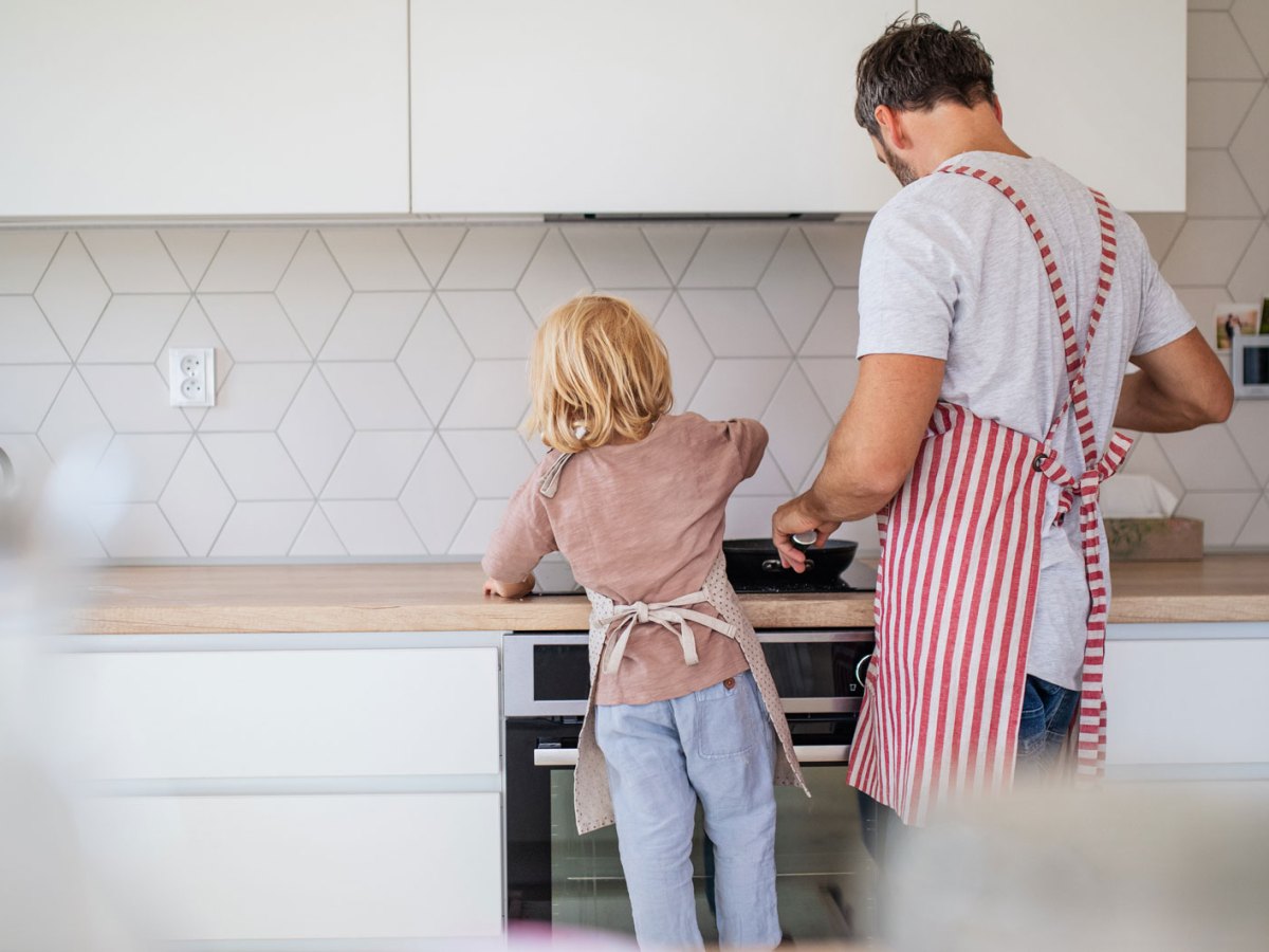Vater und Kind stehen am Herd und kochen. Man sieht beide von hinten an der Küchenzeile stehen.