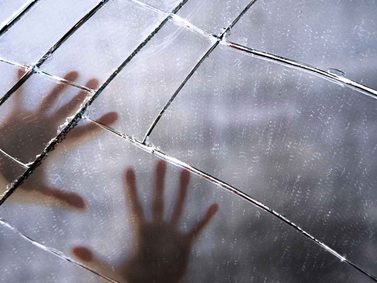 Eine unscharfe Silhouette von zwei Händen, die gegen eine zerbrochene Glasscheibe gedrückt werden, mit sichtbaren Rissen und Lichtreflexen.
