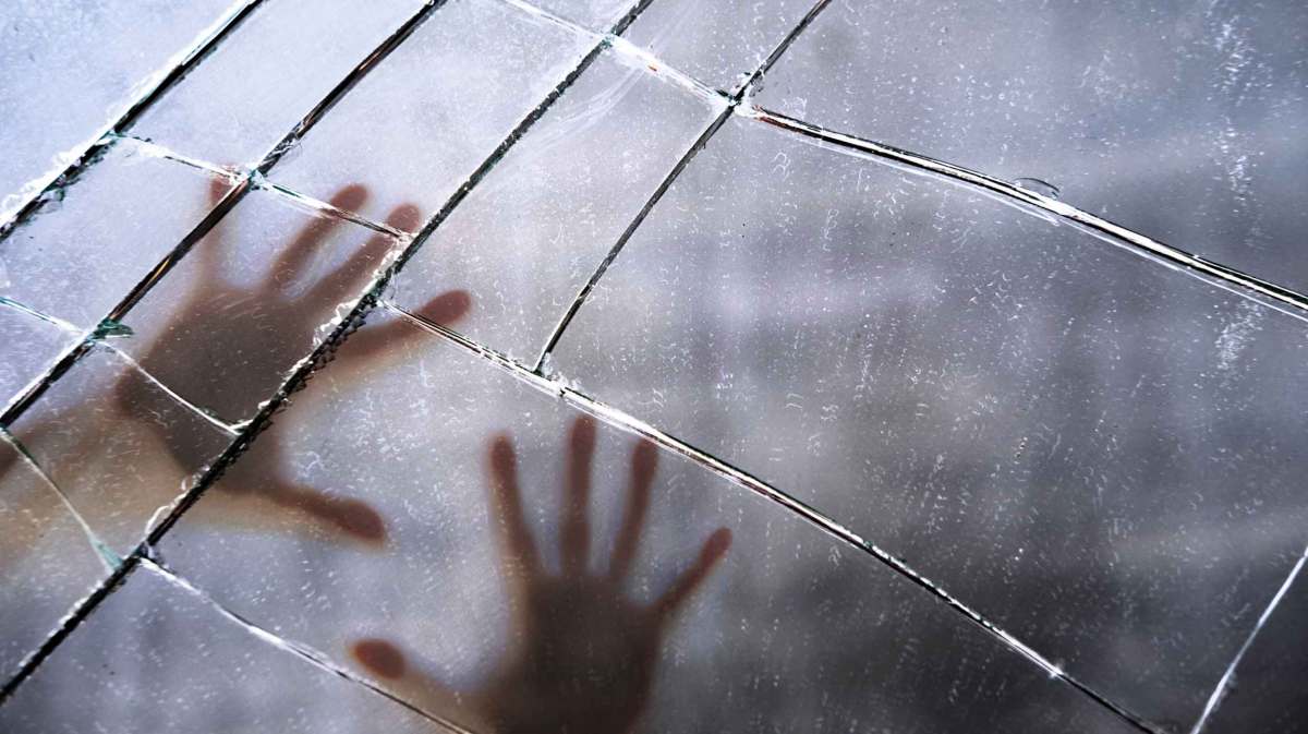 Eine unscharfe Silhouette von zwei Händen, die gegen eine zerbrochene Glasscheibe gedrückt werden, mit sichtbaren Rissen und Lichtreflexen.
