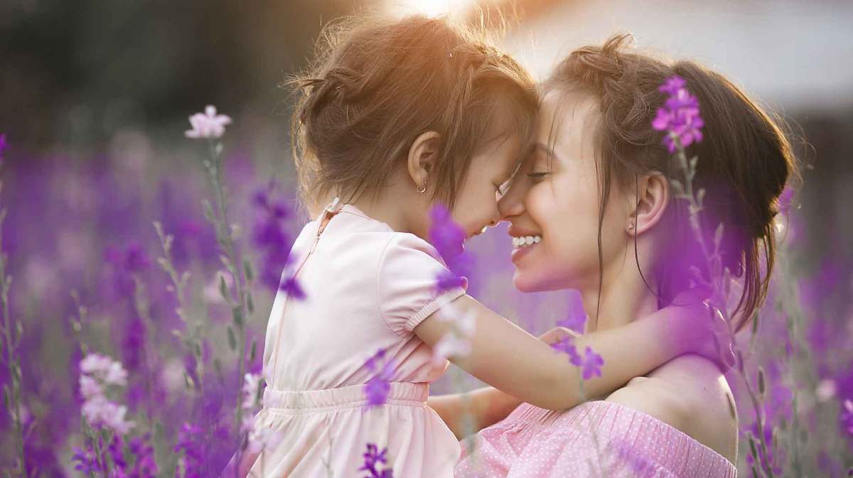 Brünette Frau hält in einem Lavendelfeld ihr Kind auf dem Arm und lächelt es an.