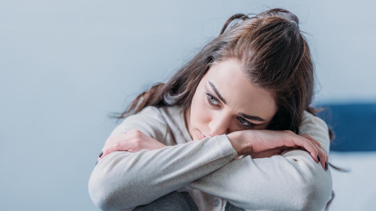 Dunkelhaarige junge Frau sitzt mit verschränkten Armen und traurigem Blick auf einem Stuhl.