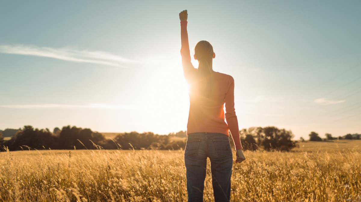 Frau auf einem Feld im Sonnenaufgang, die ihre rechte Hand in die Luft hält