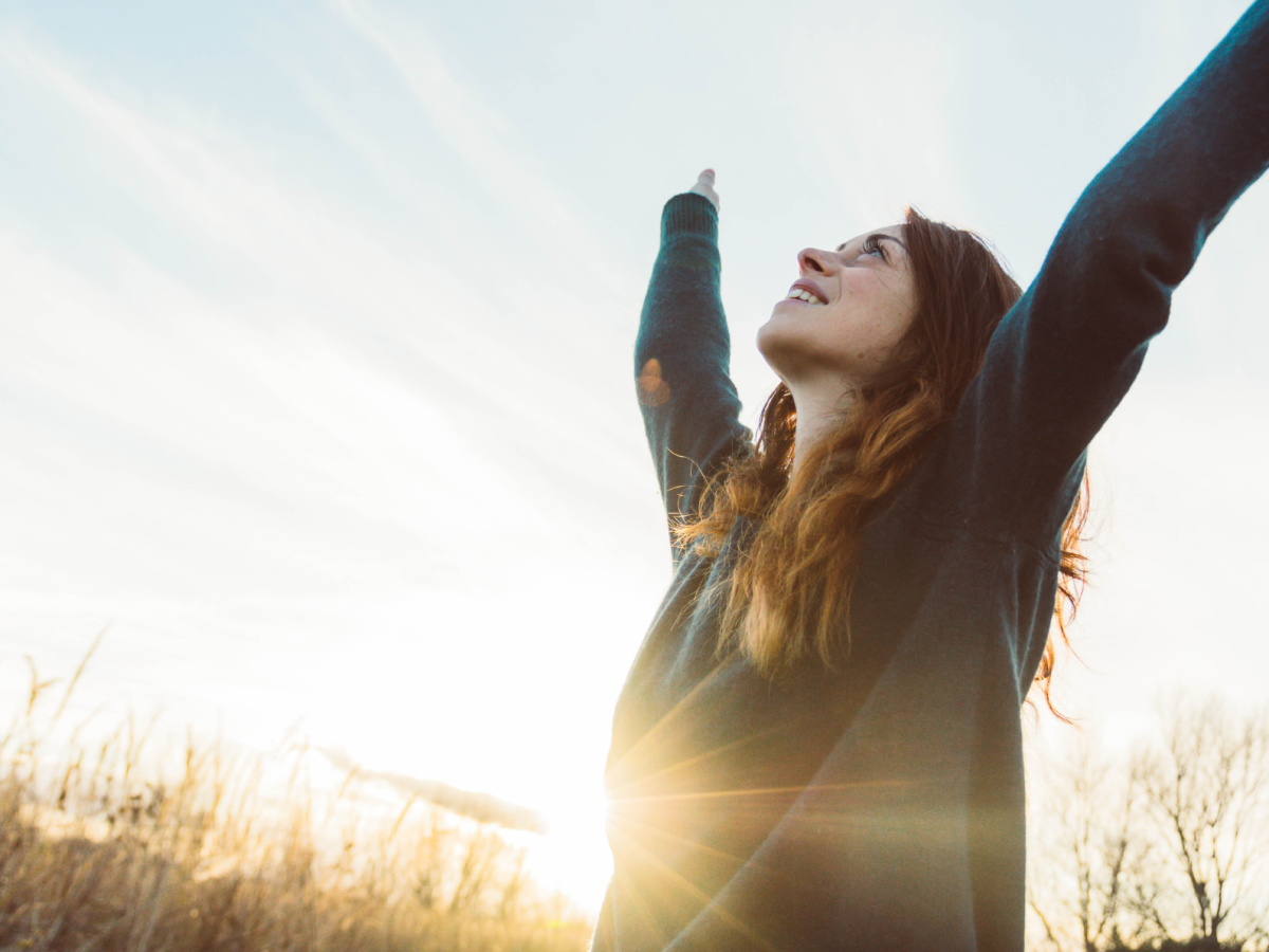 Frau in einem Feld, die ihre Arme hoch in die Luft hebt und dabei l&auml;chelt