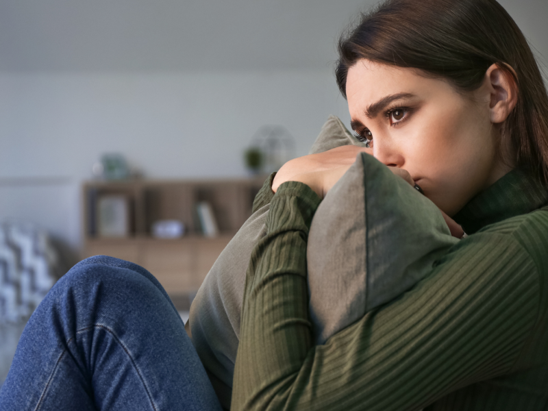Dunkelhaarige junge Frau sitzt mit traurigem Blick und einem Kissen in der Hand auf dem Sofa