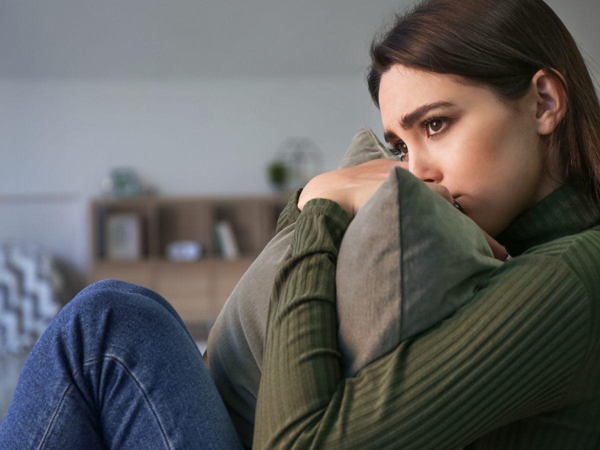 Dunkelhaarige junge Frau sitzt mit traurigem Blick und einem Kissen in der Hand auf dem Sofa