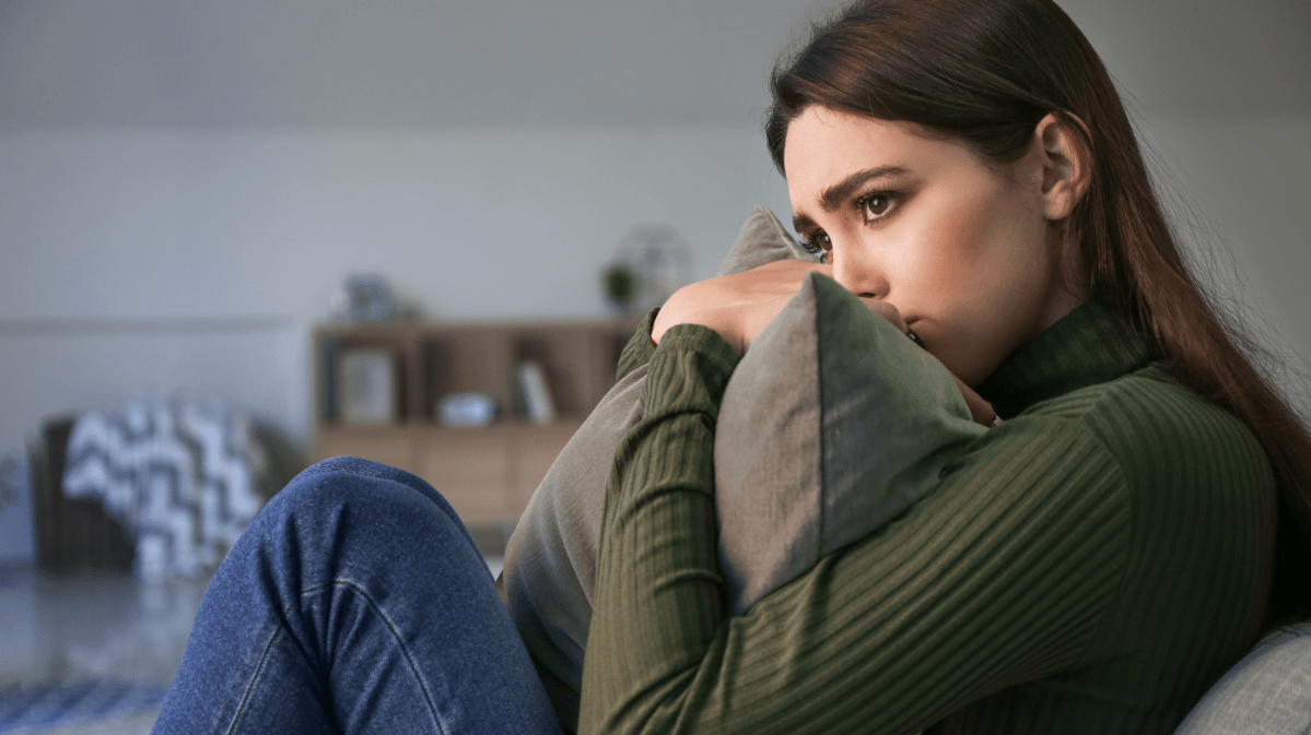 Dunkelhaarige junge Frau sitzt mit traurigem Blick und einem Kissen in der Hand auf dem Sofa