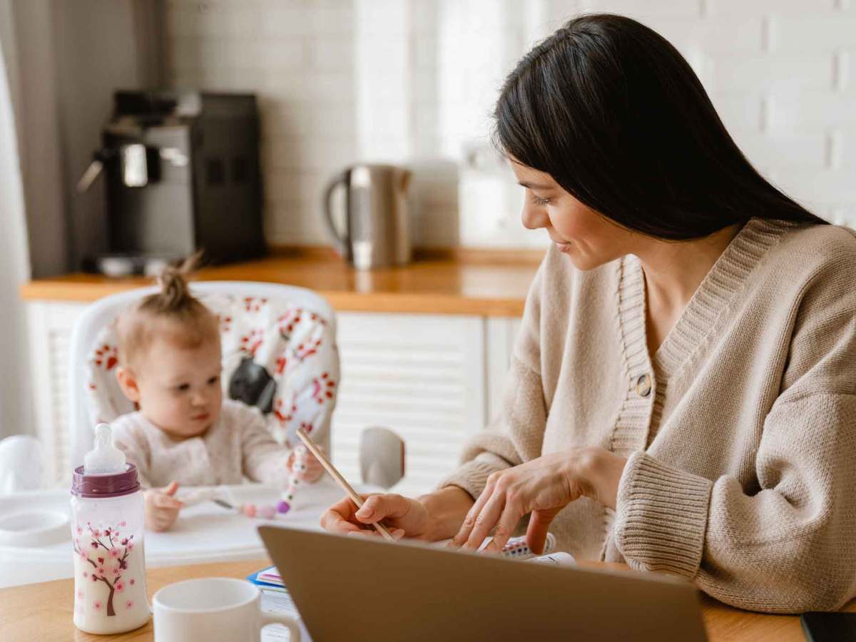Eine junge Mutter mit dunklen Haaren arbeitet konzentriert am Laptop, während ihr Kleinkind im Hochstuhl daneben spielt.
