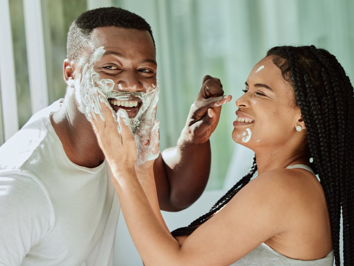 Junges Paar lacht verspielt im Badezimmer, beide schmieren sich Rasierschaum ins Gesicht vor einem hellen Fenster.