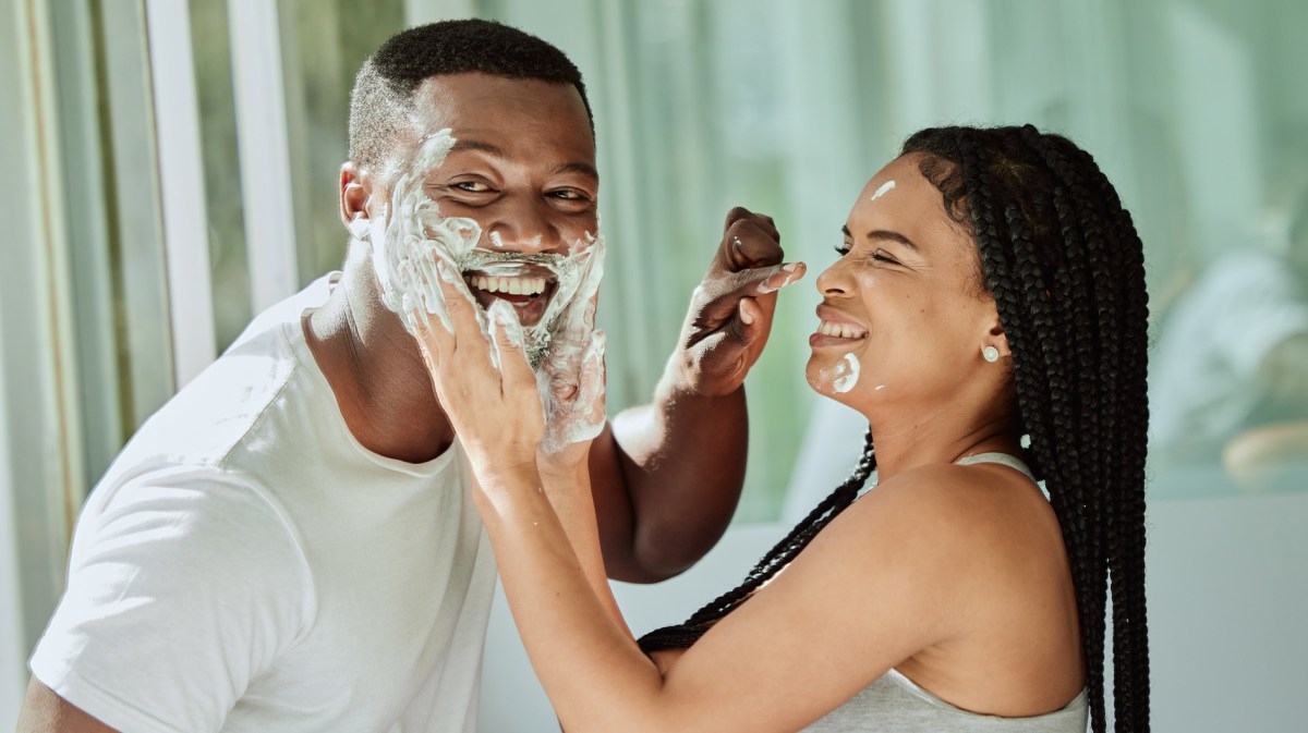 Junges Paar lacht verspielt im Badezimmer, beide schmieren sich Rasierschaum ins Gesicht vor einem hellen Fenster.