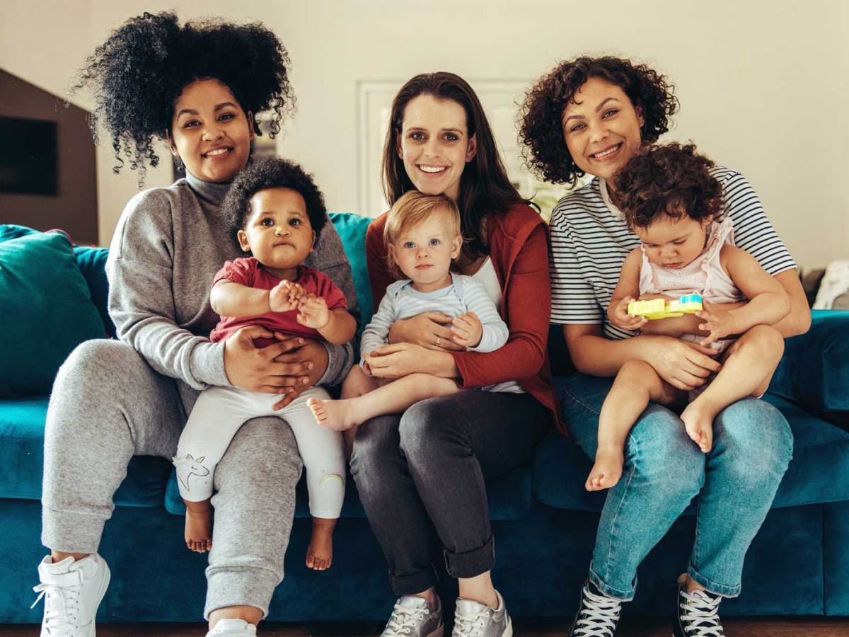 Drei Mamas haben ihre Kleinkinder auf dem Schoß, sitzen auf einem blauen Sofa und strahlen in die Kamera.