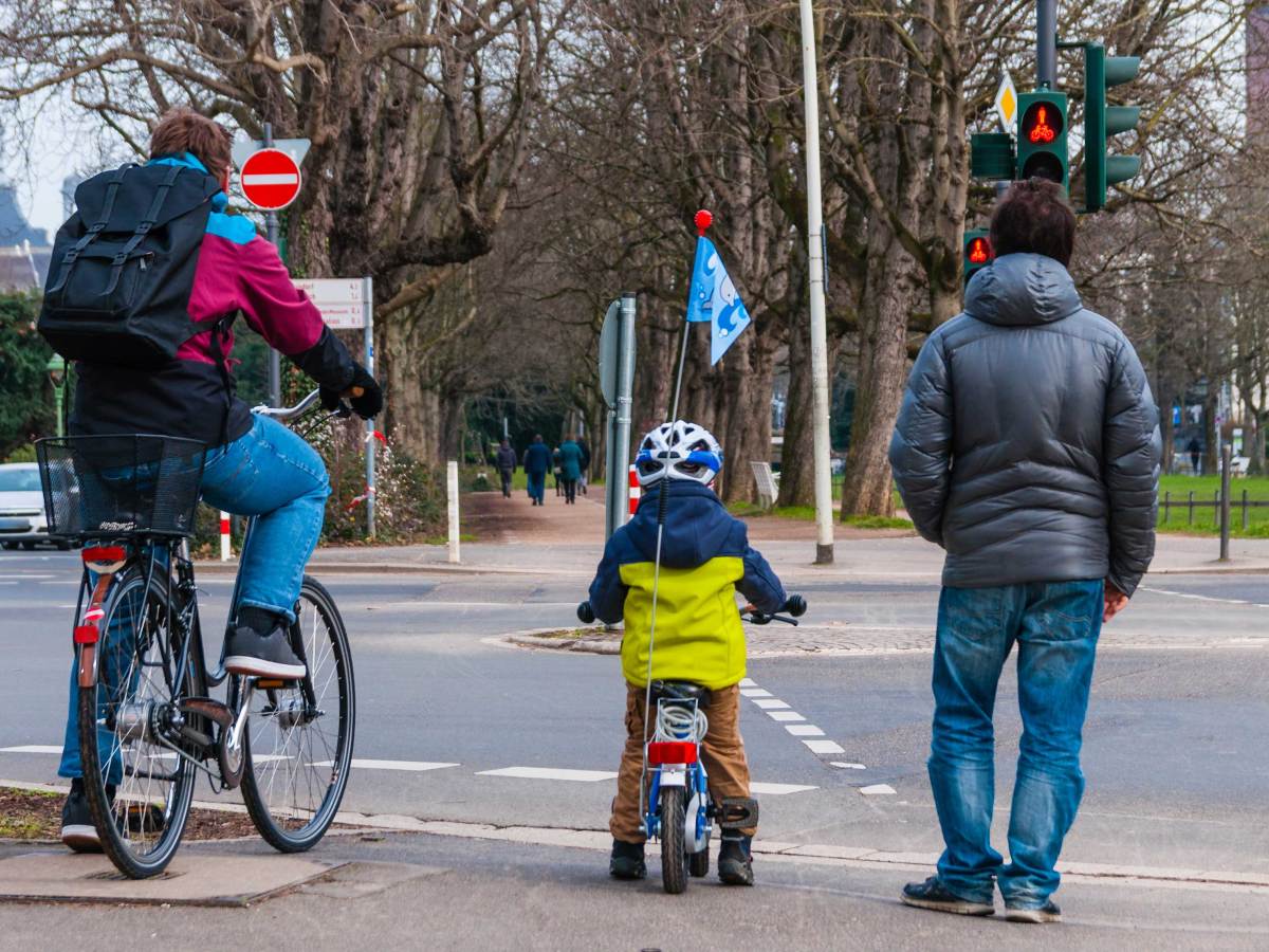 Vater und ca. 4-jähriges Kind stehen auf ihren Fahrrädern sitzend an der Ampel und warten auf Grün.