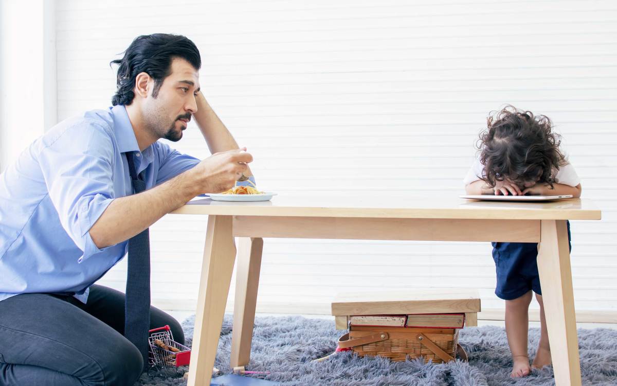 Vater schaut frustriert auf sein kleines Kind, das bockig den Kopf auf den Tisch gelegt hat, statt zu essen.