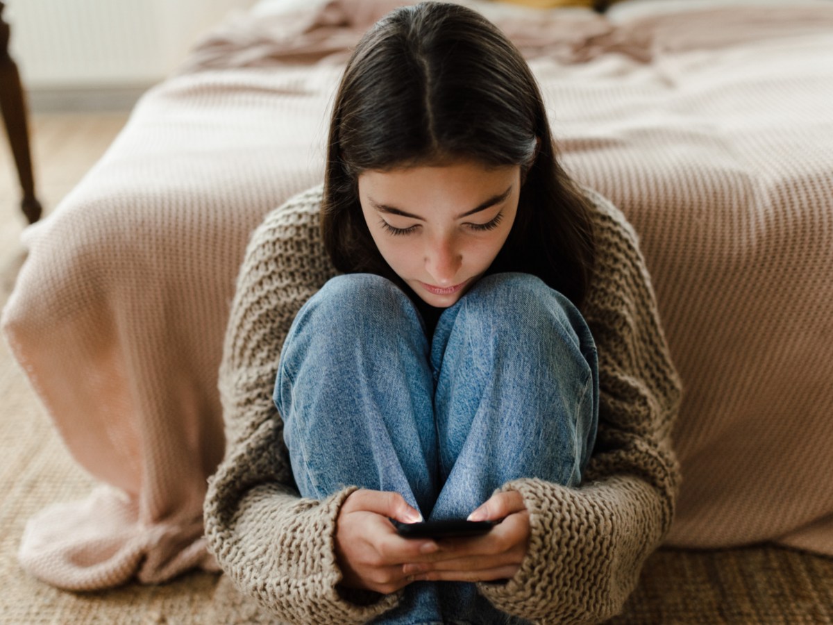 Frau, die vor ihrem Bett sitzt und sch&uuml;chtern auf ihr Handy starrt