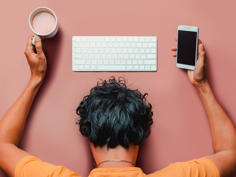 Mann vor rosa Hintergrund, der ein Handy und einen Kaffee in der Hand hält, während sein Kopf vor der Tastatur auf dem Boden liegt