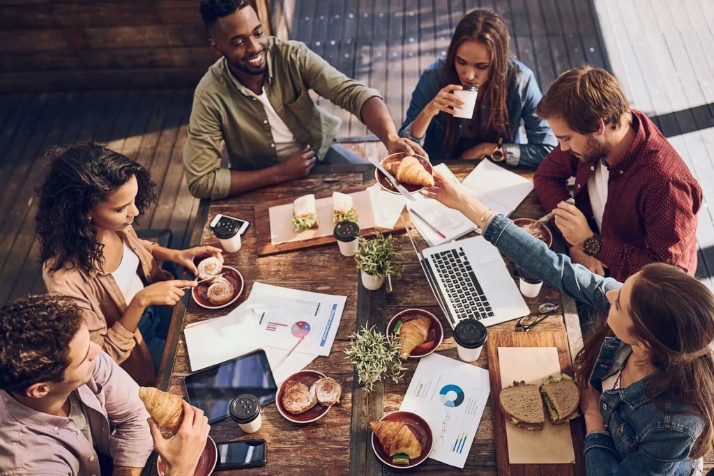 Frauen und Männer sitzen an einem Tisch und arbeiten und essen gemeinsam