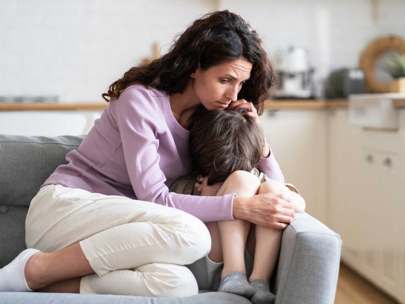 Mutter und Sohn sitzen auf dem Sofa. Er versteckt seinen Kopf zwischen seinen Beinen und seine Mutter tröstet ihn und hält ihn fest im Arm.