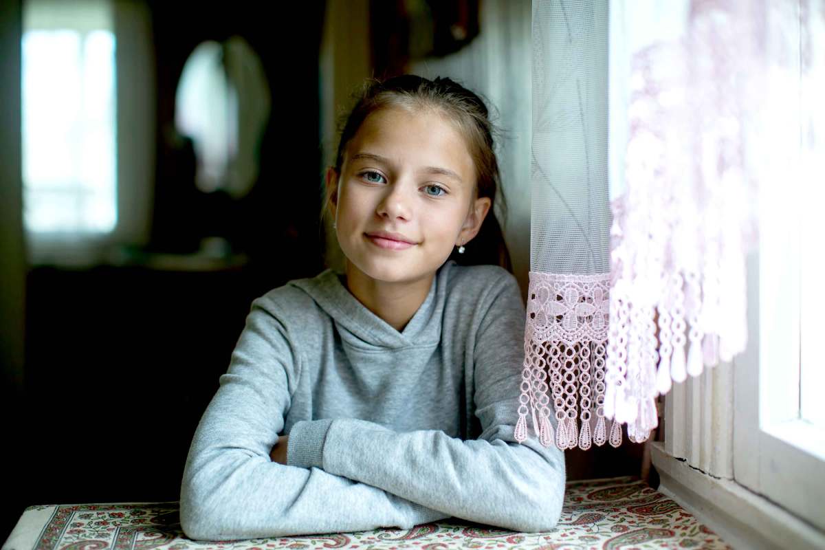 Mädchen, ca. 12 Jahre sitzt mit verschränkten Armen aber einem freundlichen Lächeln am Tisch.