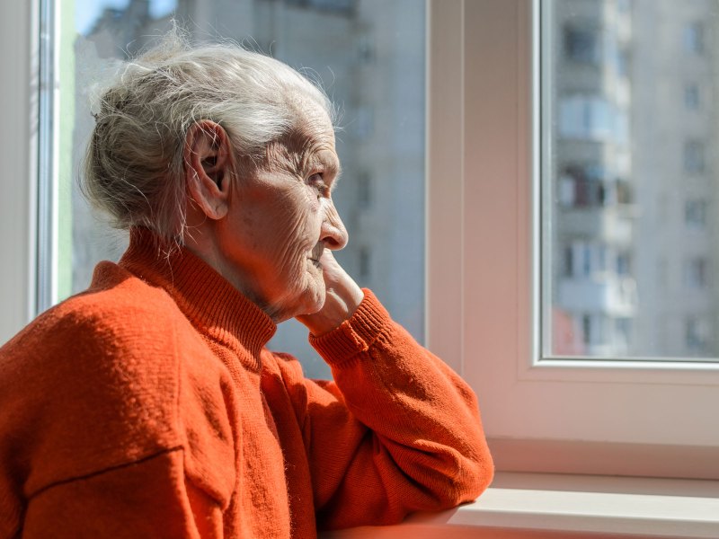 alte Frau sitzt in einem orangefarbenen Pullover am Fenster