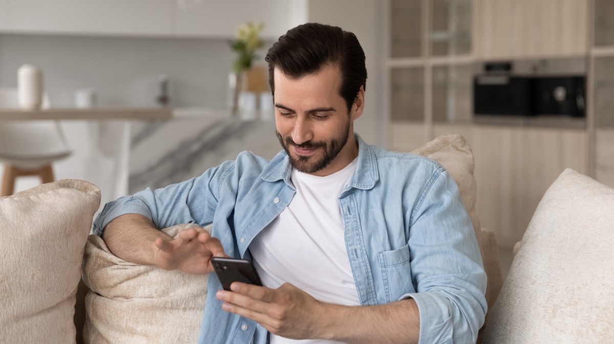 Mann der auf der Couch sitzt und sein Handy in der Hand hält, während er lächelt