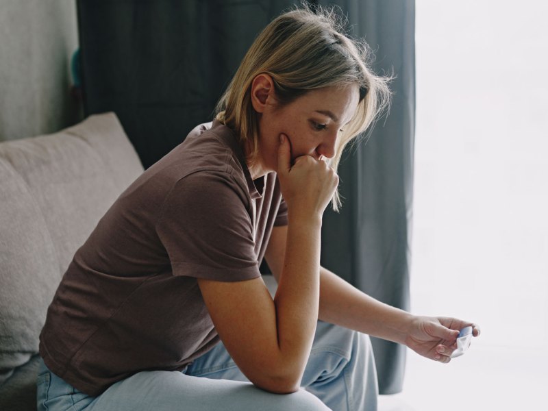 Frau sitzt im Wohnzimmer und schaut erschrocken auf einen Schwangerschaftstest in ihrer Hand.