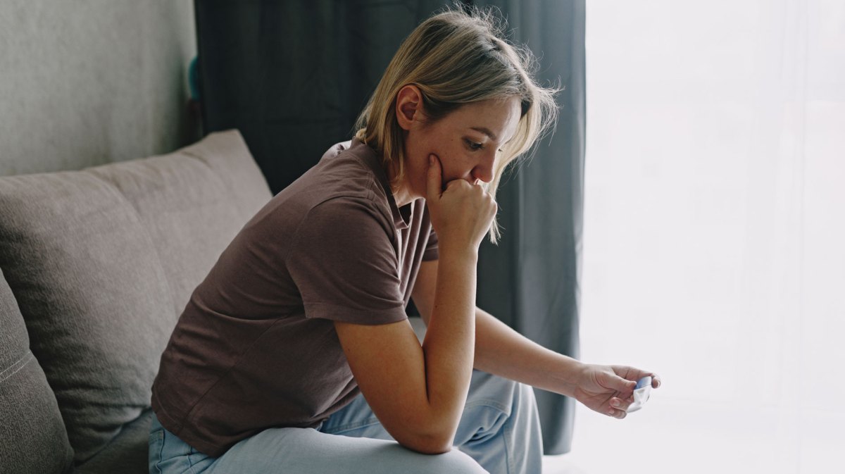 Frau sitzt im Wohnzimmer und schaut erschrocken auf einen Schwangerschaftstest in ihrer Hand.