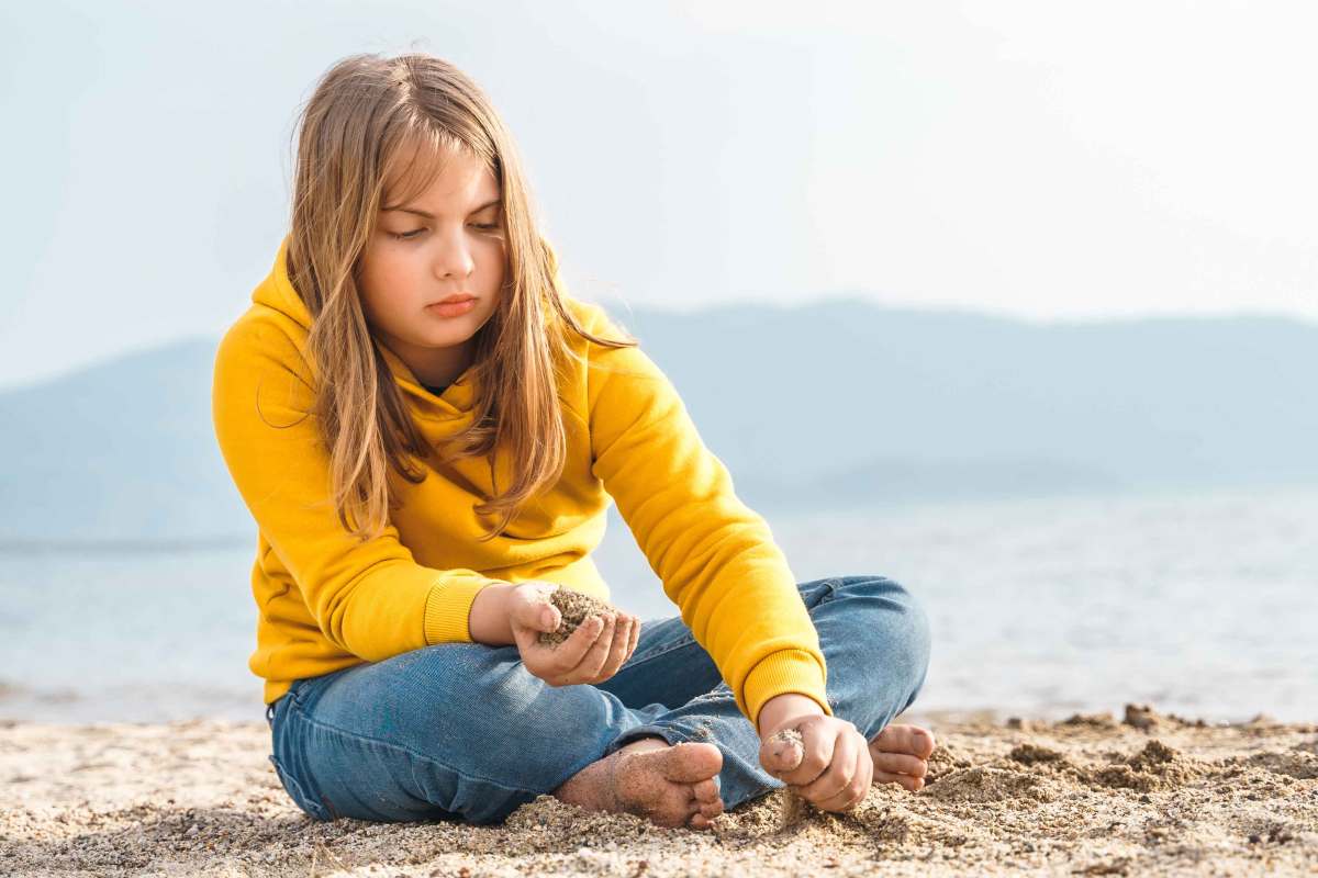 Mädchen um die 12 Jahre sitzt in Jeans und Hoodie alleine am Strand und spielt gedankenversunken mit dem Sand.