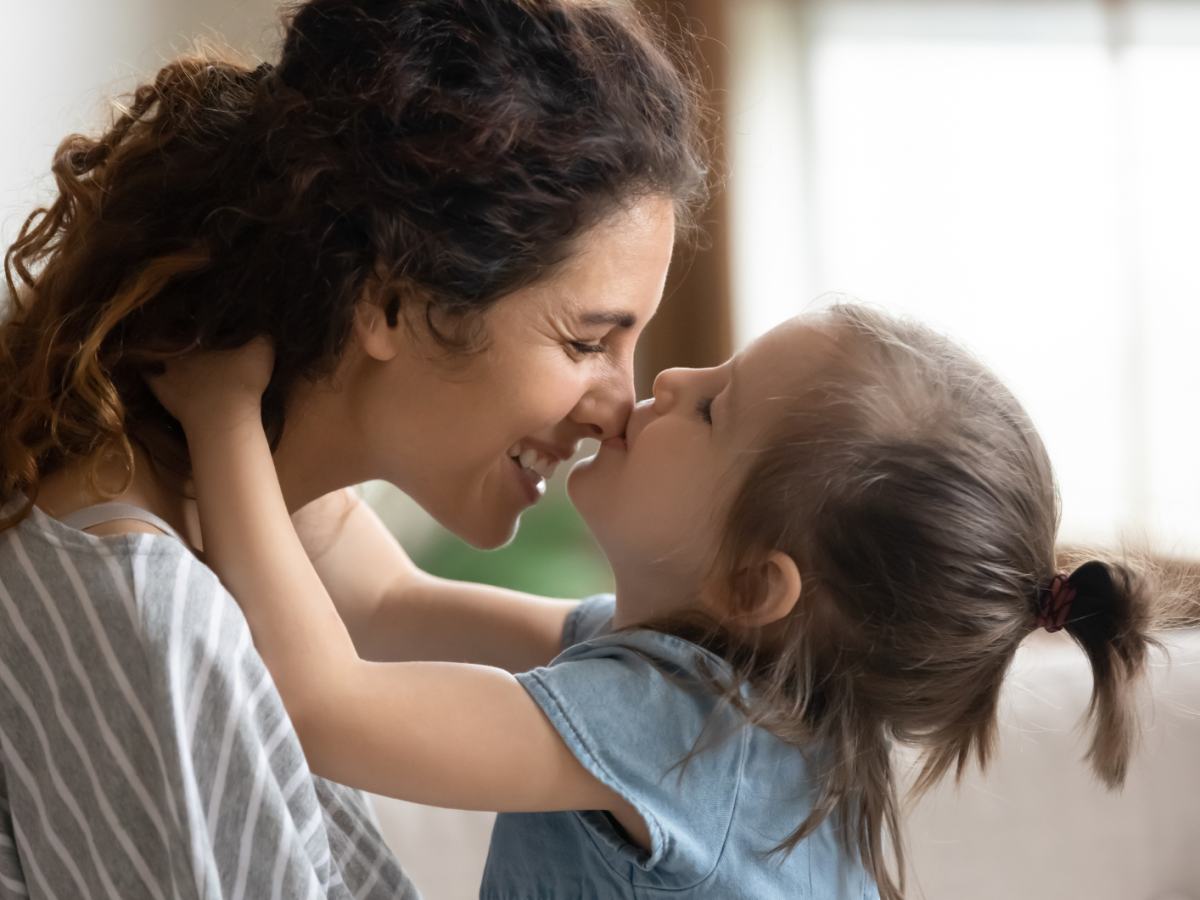 Mutter hält ihre Tochter vor sich, die sie auf die Nase küsst.