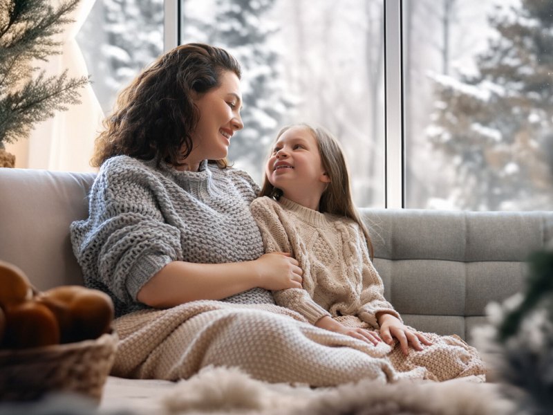 Mutter und Tochter sitzen eingekuschelt in eine Decke auf dem Sofa und unterhalten sich.