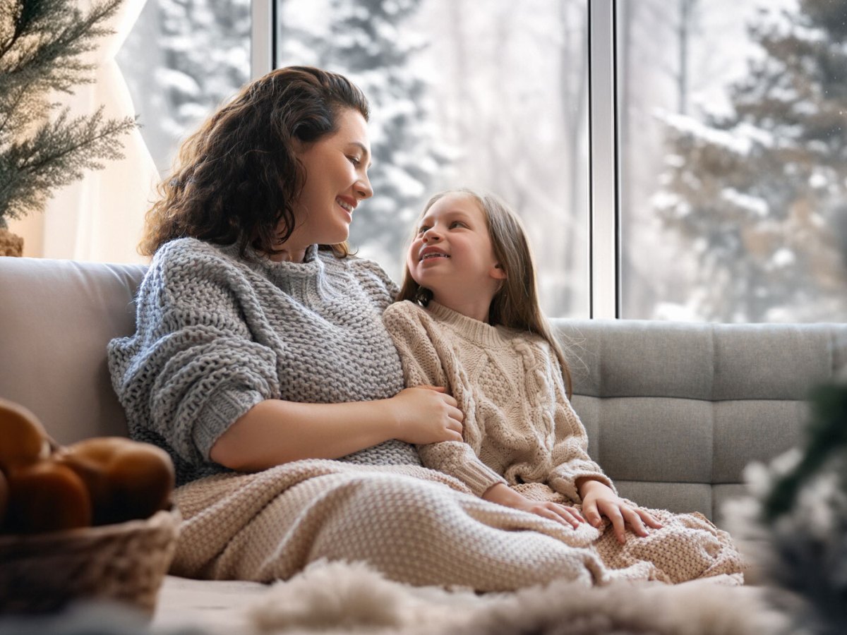 Mutter und Tochter sitzen eingekuschelt in eine Decke auf dem Sofa und unterhalten sich.