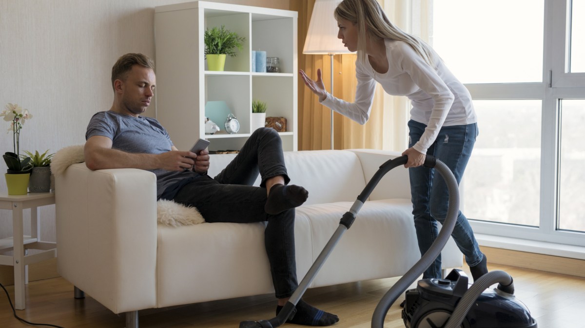 Mann sitzt auf Sofa während Frau staubsaugt.