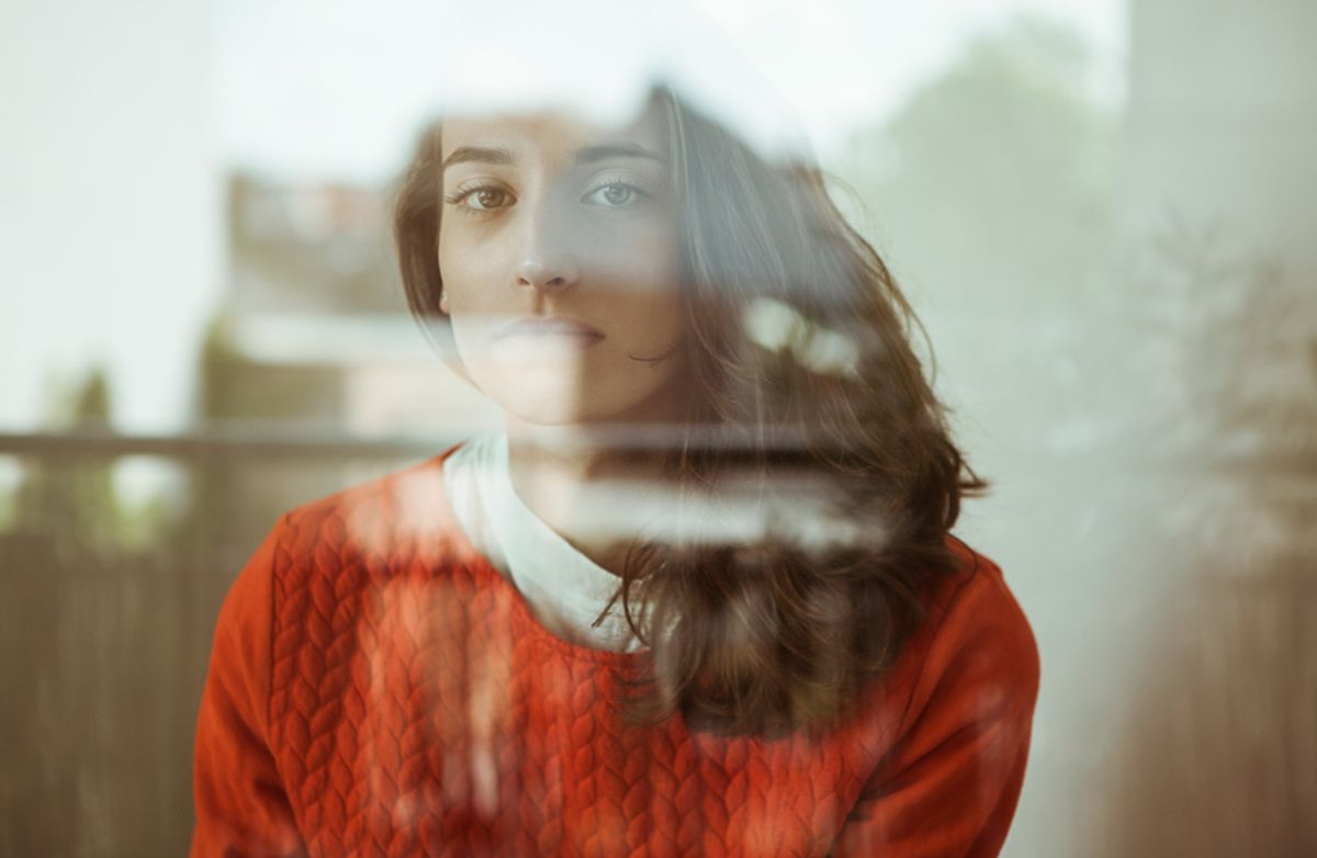 Frau schaut ernst durch ein spiegelndes Fenster.