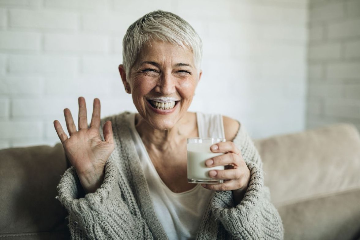 &Auml;ltere Frau mit grauem Haar lacht fr&ouml;hlich mit Buttermilch-Bart in die Kamera.