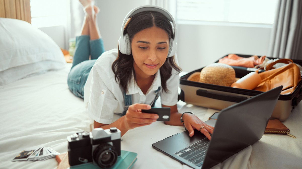 Frau mit Kopfhörern liegt auf dem Bett, mit Laptop und gepacktem Koffer. Hält Kreditkarte in der Hand.